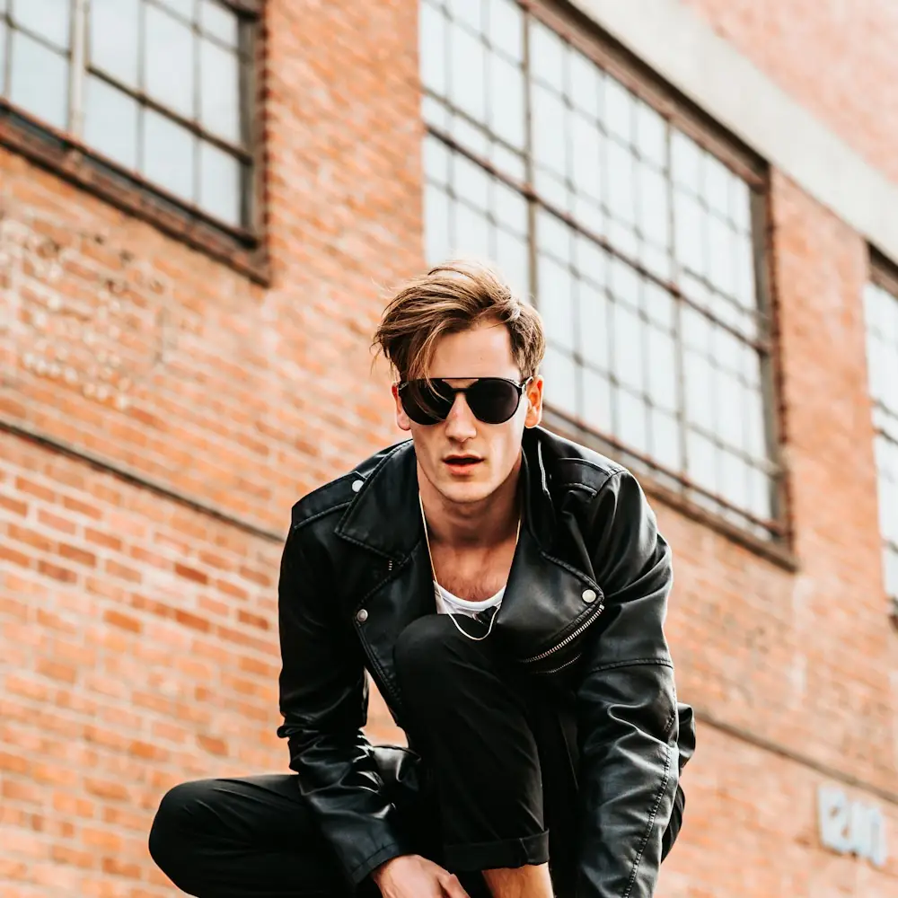 An urban rider in a leather jacket and sunglasses in front of a brick warehouse wall